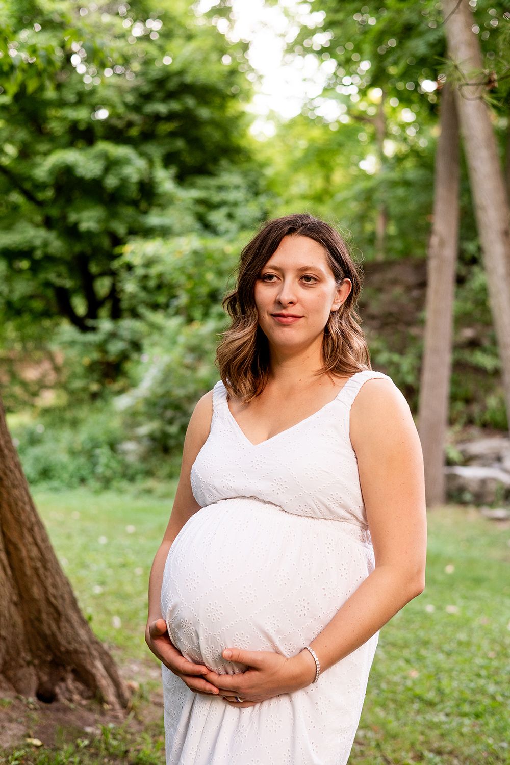 Woman in a white dress cradles her pregnant belly while standing in a lush garden setting.
