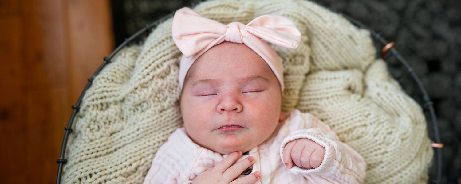 A peacefully sleeping baby wearing a pink outfit and bow headband rests on a cream-colored knit blanket.