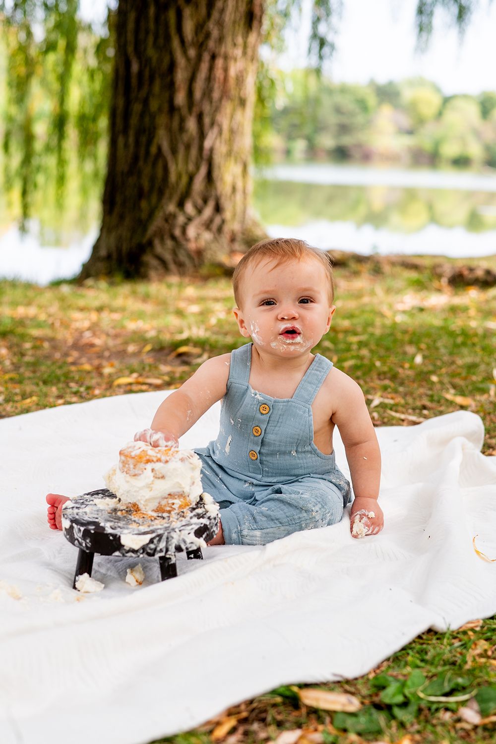 Small child wearing denim overalls sits on white blanket under willow trees beside lake on autumn day.
