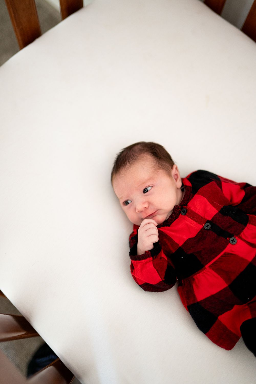 A baby wearing red and black buffalo plaid sleepwear lies on white bedding looking at the camera.
