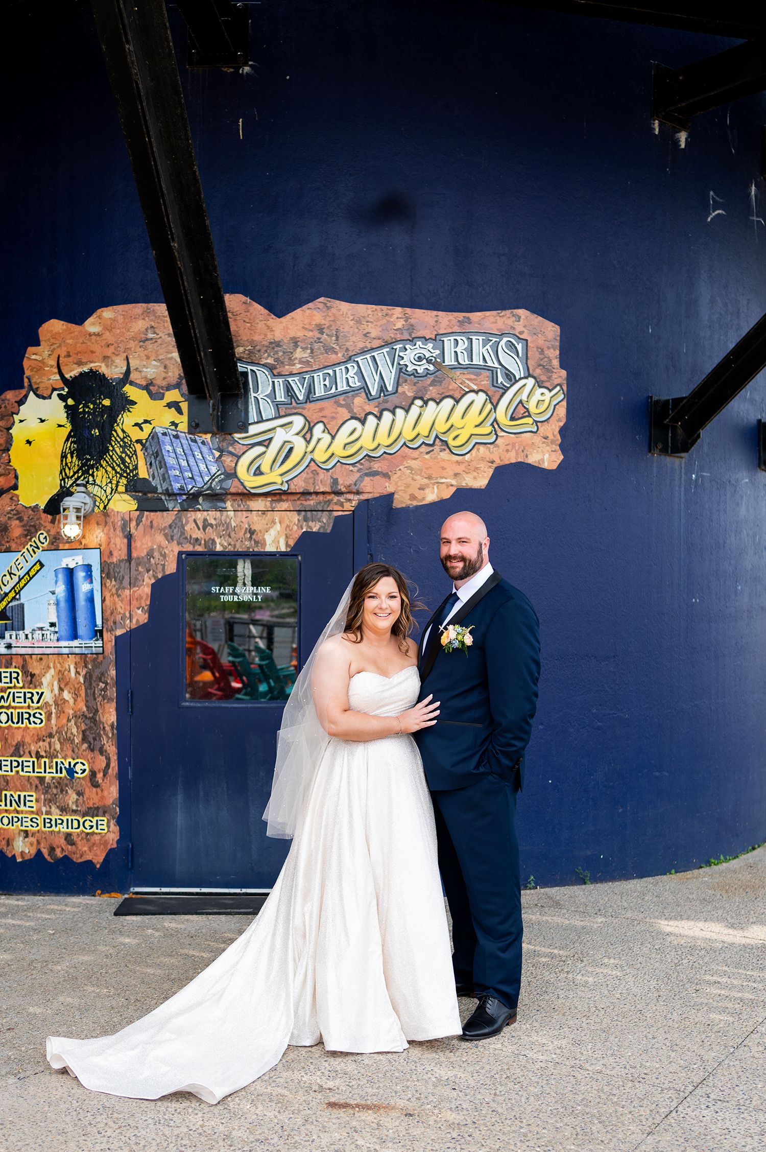 A wedding couple poses in front of a colorful painted wall mural at a riverfront venue.