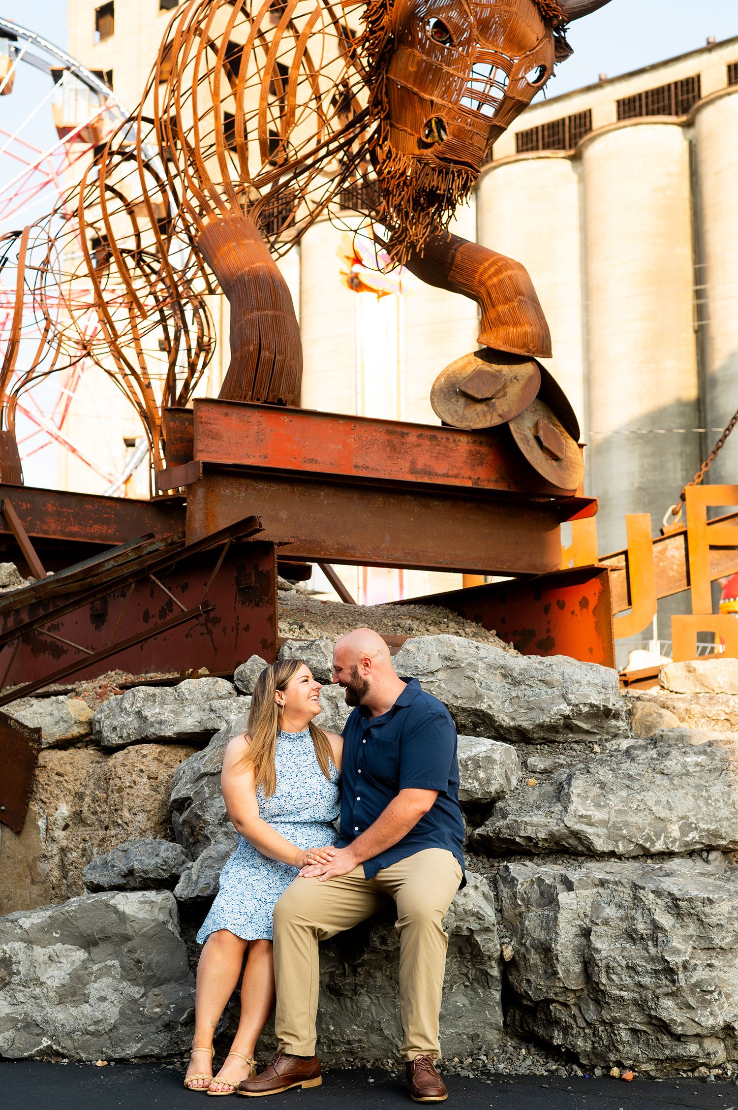 Couple sitting close together on stone wall beneath large rusty metal lion sculpture outdoors.
