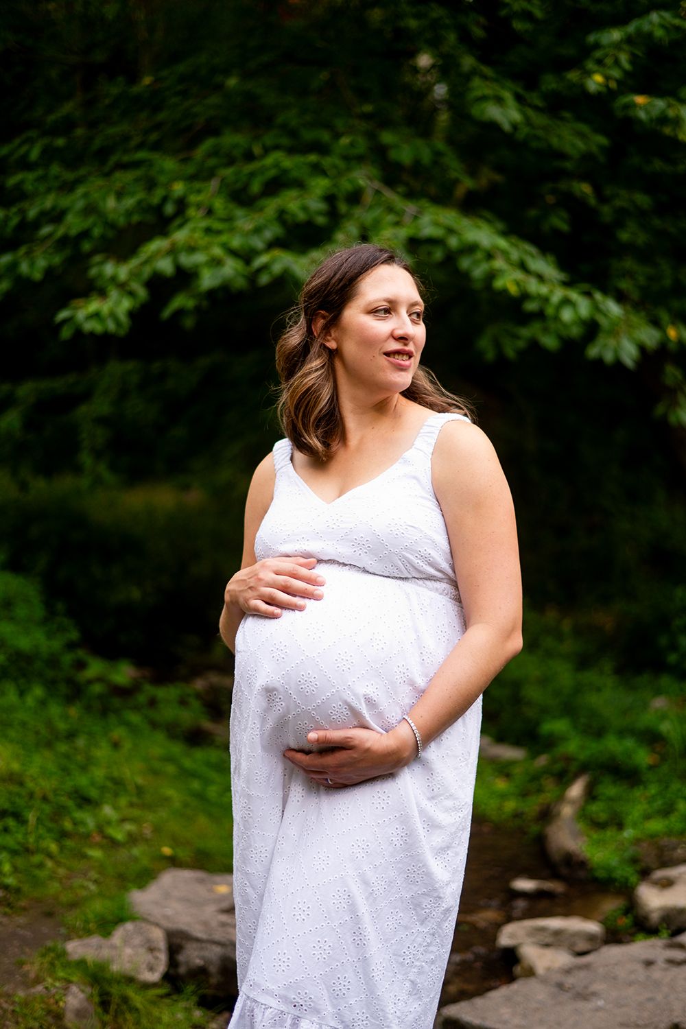 Person in a white maxi dress poses outdoors during a maternity photo session with greenery in the background.