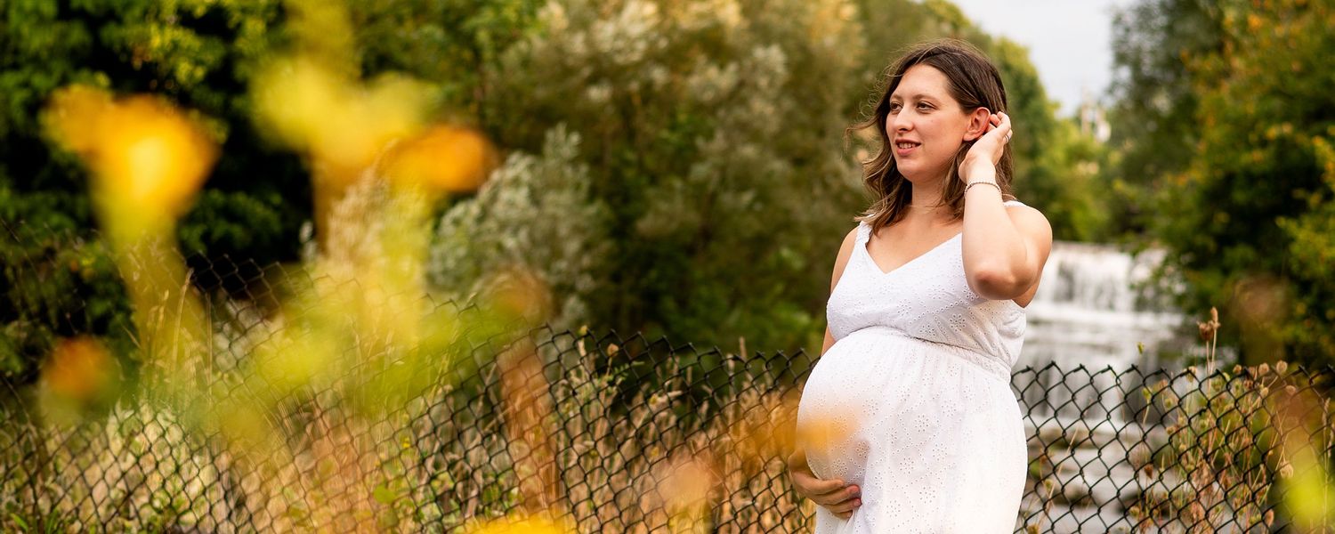 Expectant person in white dress poses outdoors in golden sunlight with bokeh flowers in foreground.