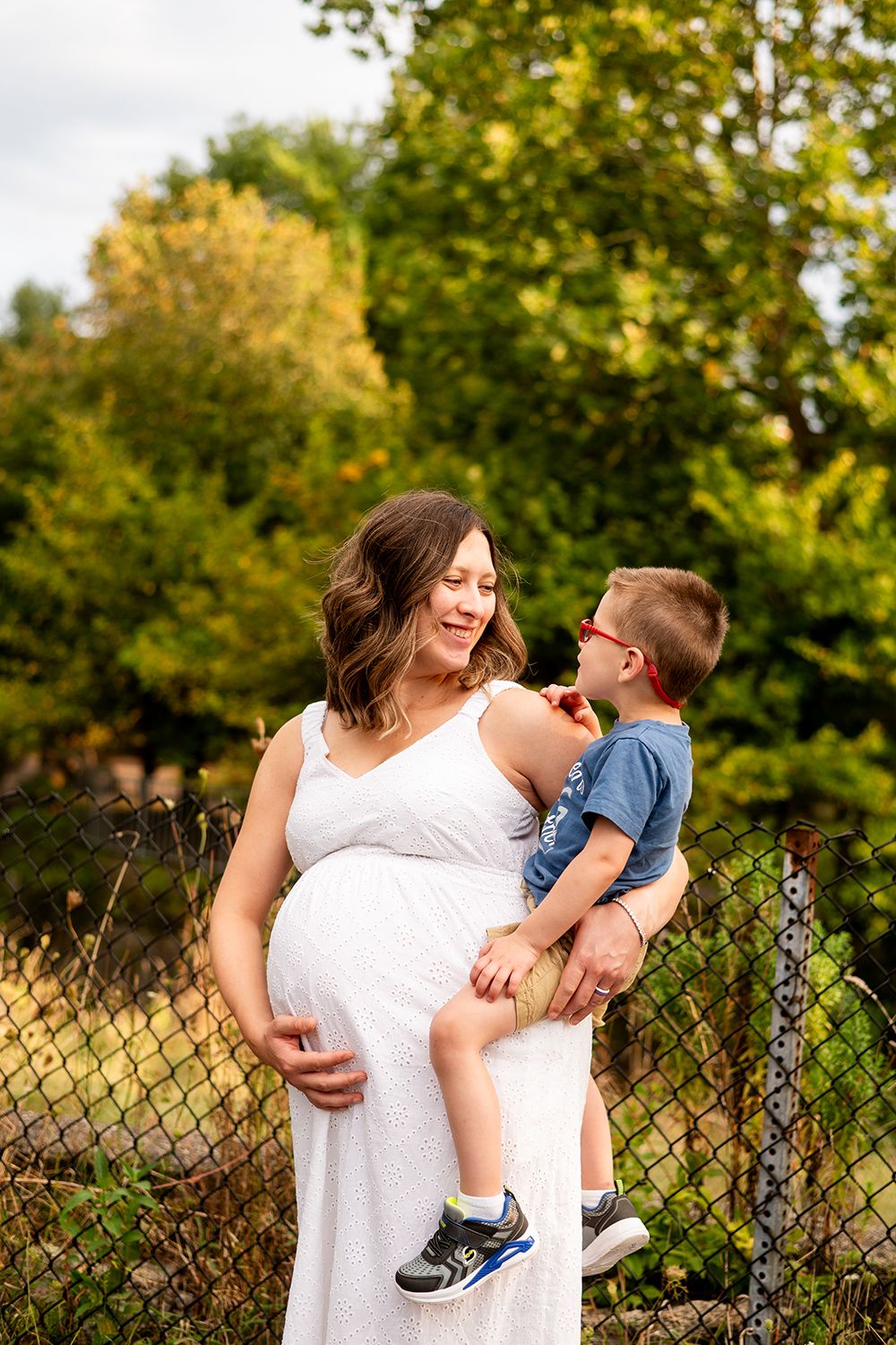 A pregnant woman in a white dress holds a young child outdoors on a sunny day with trees in the background.