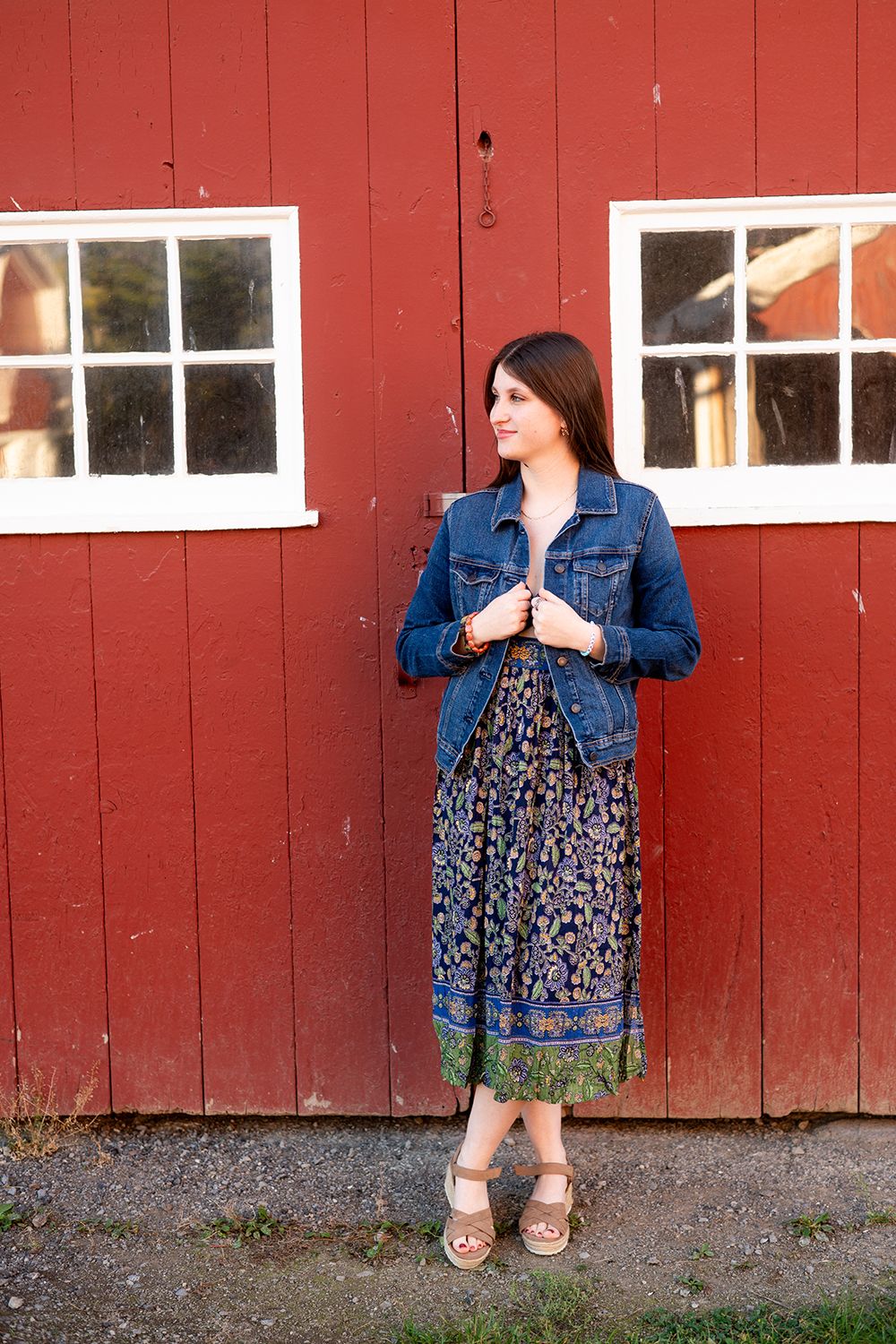 Person wearing a denim jacket and floral dress poses against a red barn wall with white windows.