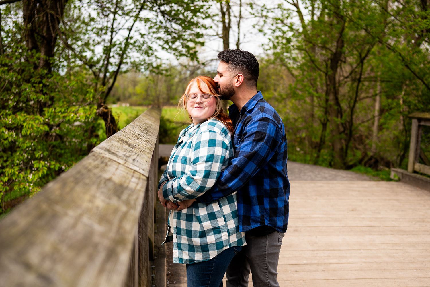 A couple wearing plaid and denim embrace on a wooden bridge surrounded by lush spring greenery.