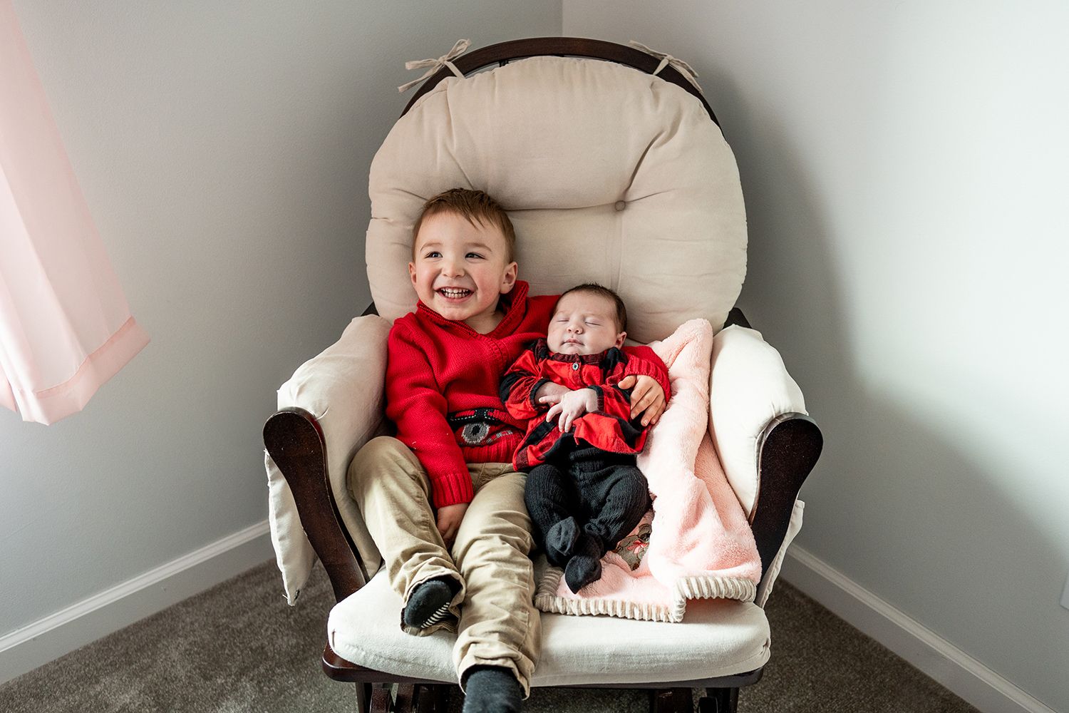 Two children sitting together in a cream-colored glider chair, wearing matching red holiday outfits.