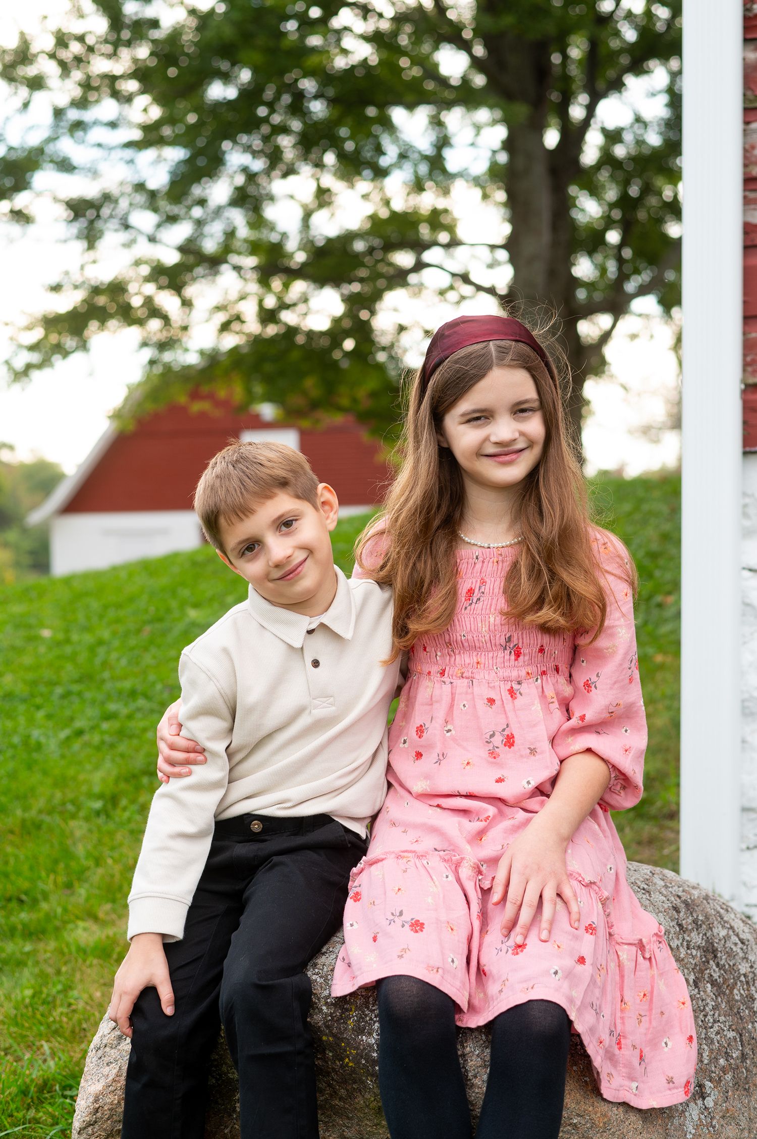 Two children, a boy in a cream polo and a girl in a pink floral dress, sit together on a rock outdoors.