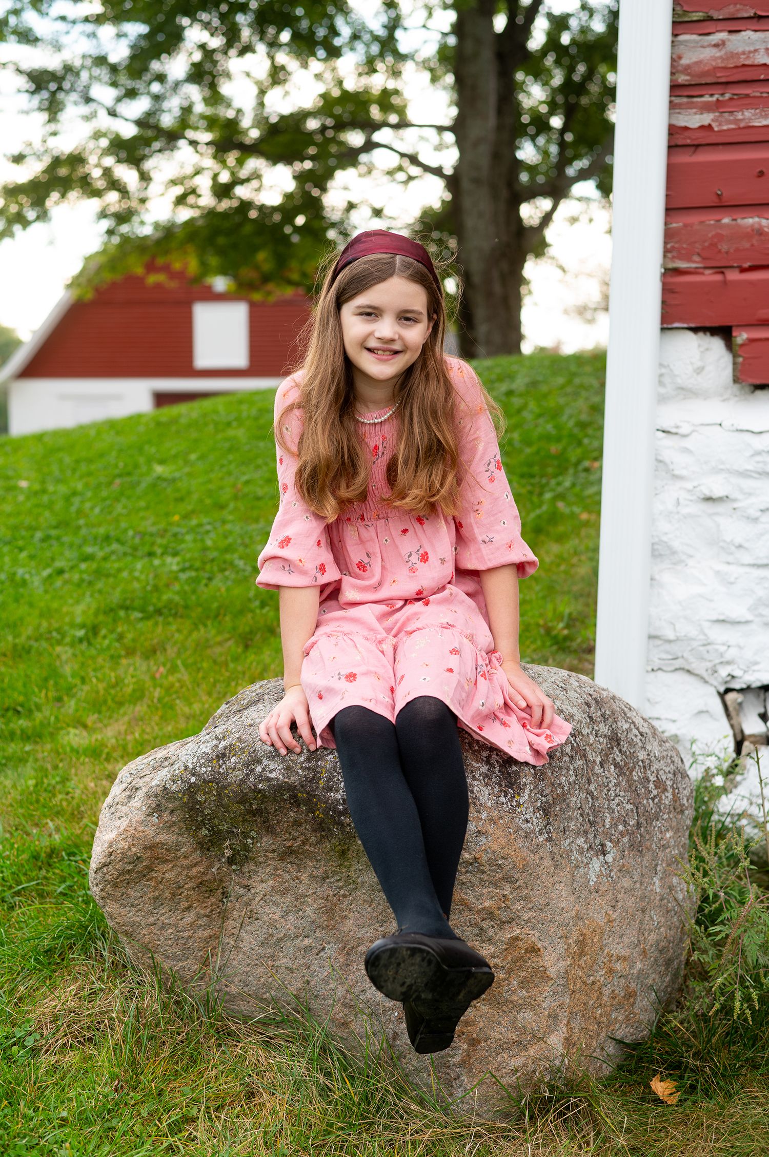Young girl in pink dress and black tights sitting on a large rock outdoors near a red barn.