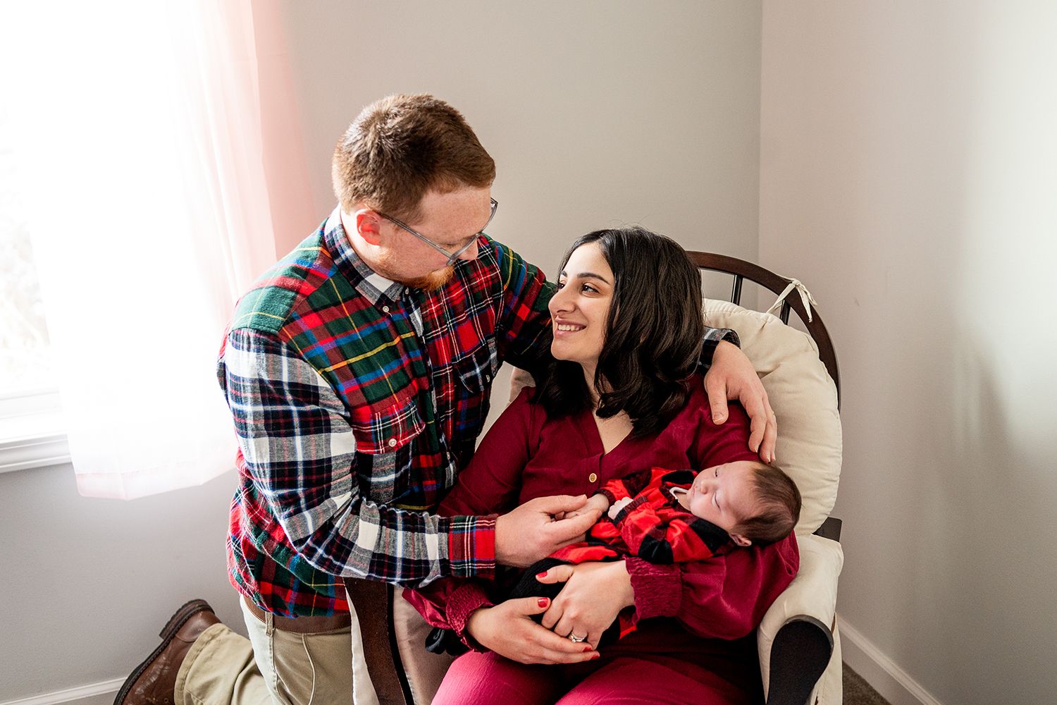 A couple wearing festive red and plaid clothing share a tender moment in a cozy white nursery.