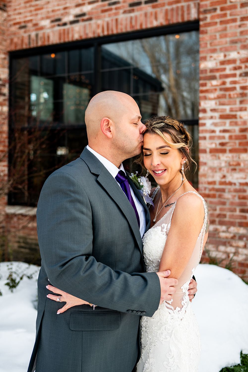 An intimate wedding portrait against a rustic brick wall with snow on the ground.