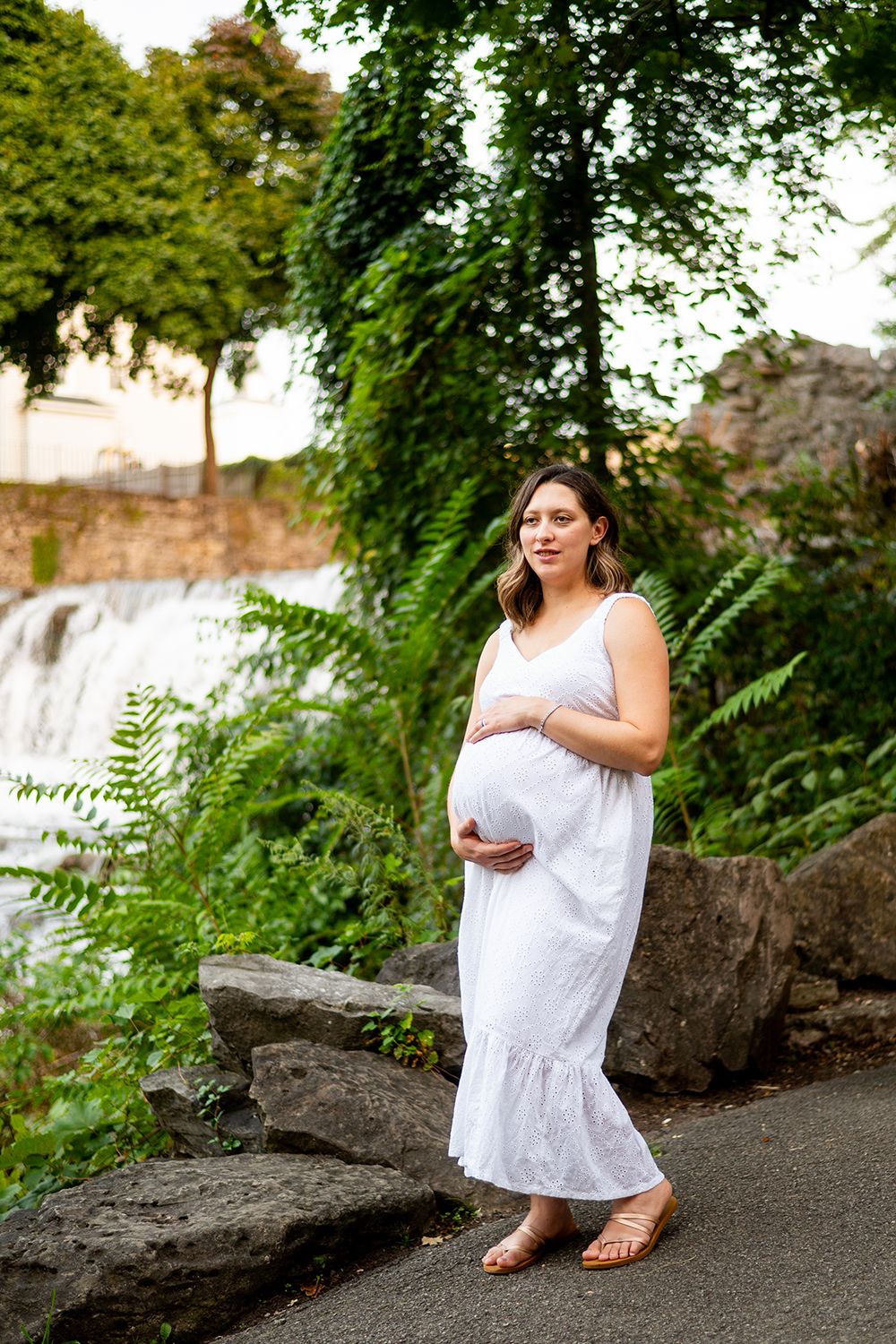 A pregnant woman in a white sundress stands near a waterfall and lush greenery on a summer day.