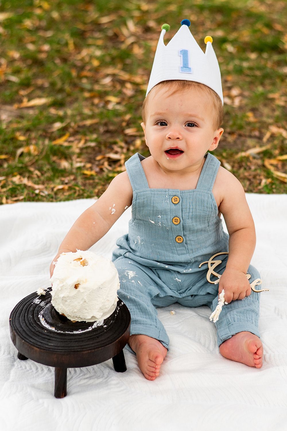 A first birthday photoshoot with white crown and denim overalls, sitting on blanket with smash cake on wooden pedestal.