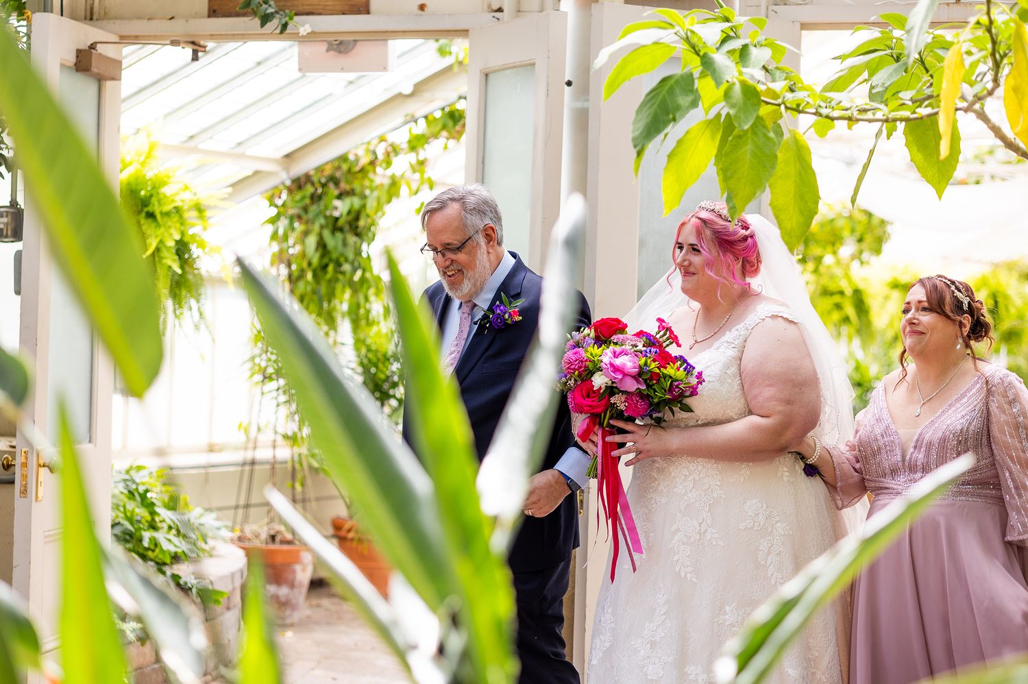 Bride with pink hair holding colorful bouquet walks with escort through lush greenhouse wedding venue.