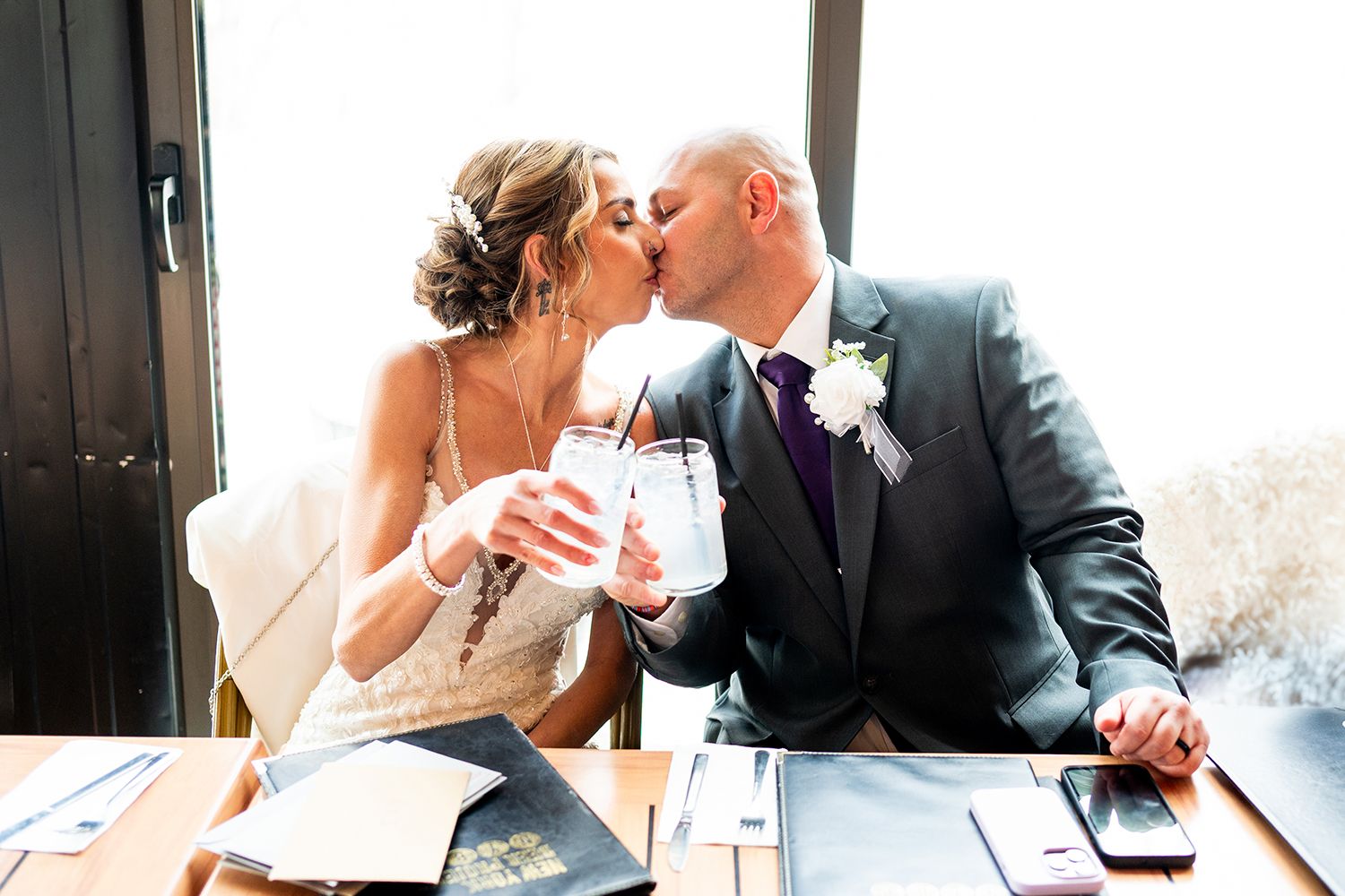 A couple in wedding attire share a romantic kiss while seated at a desk with paperwork.