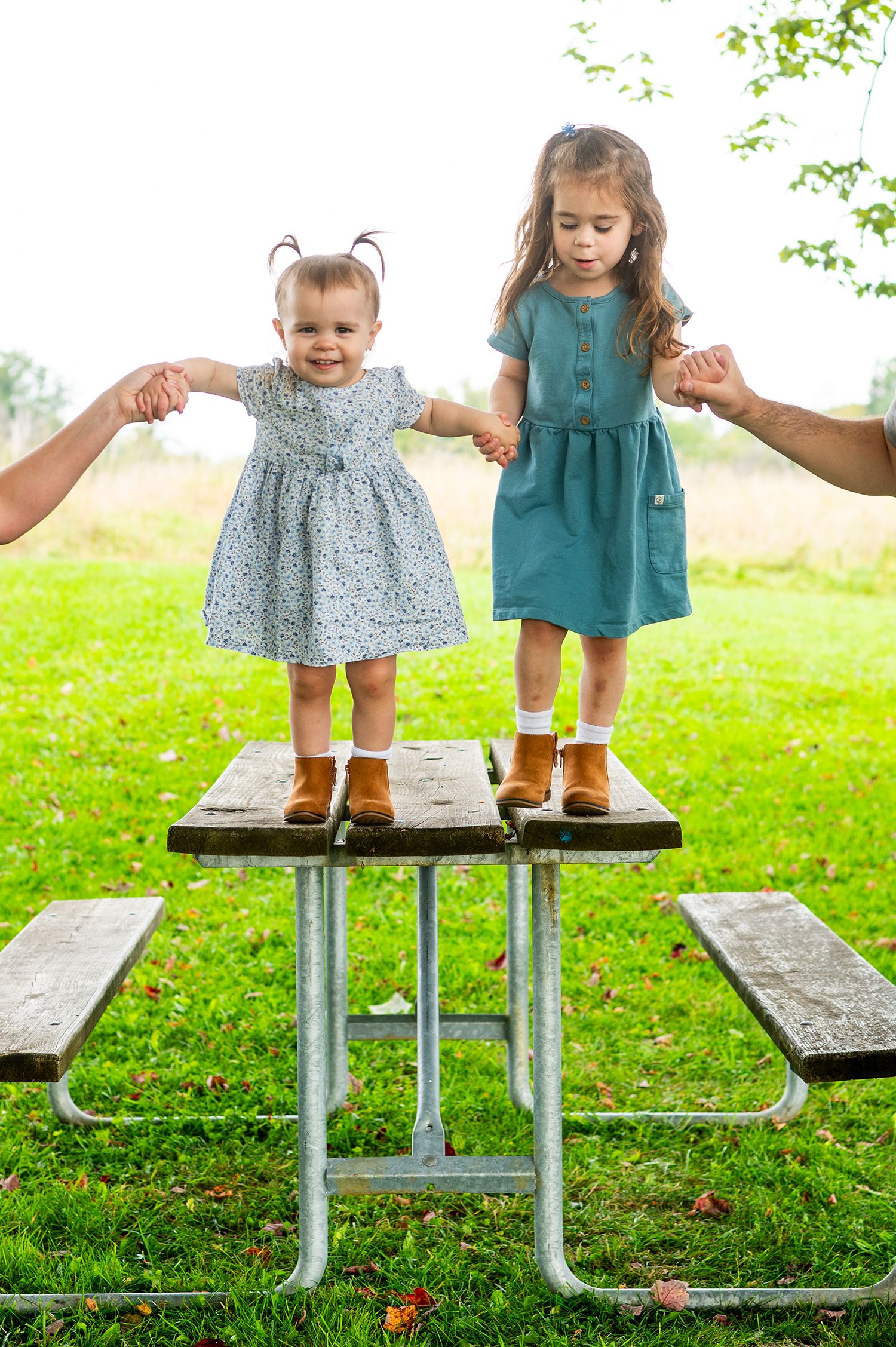 Two young girls in dresses and boots standing on a picnic table outdoors, held by adults hands.