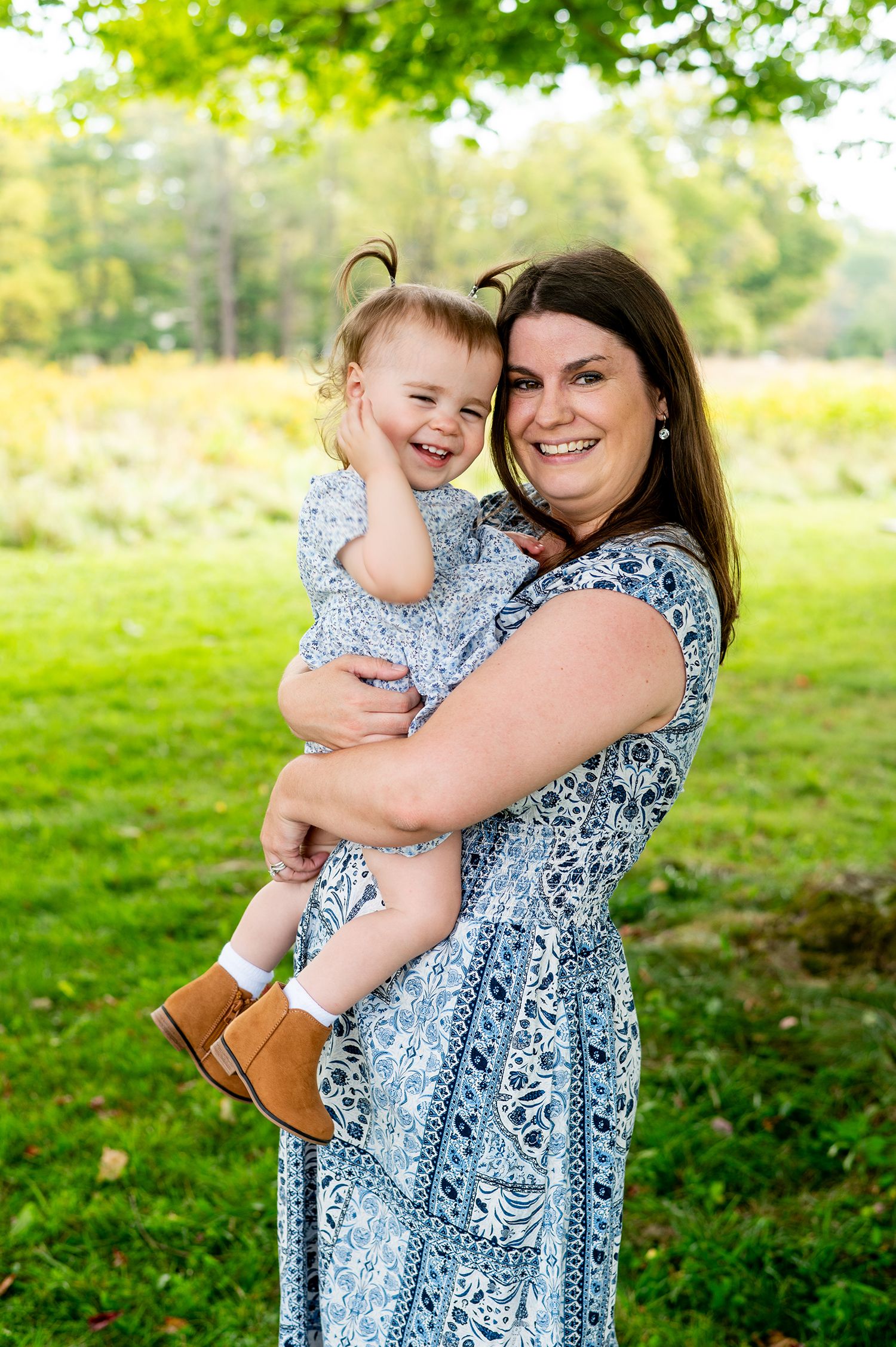 A smiling mother in a blue floral dress holds her laughing toddler daughter outdoors in a lush green park.
