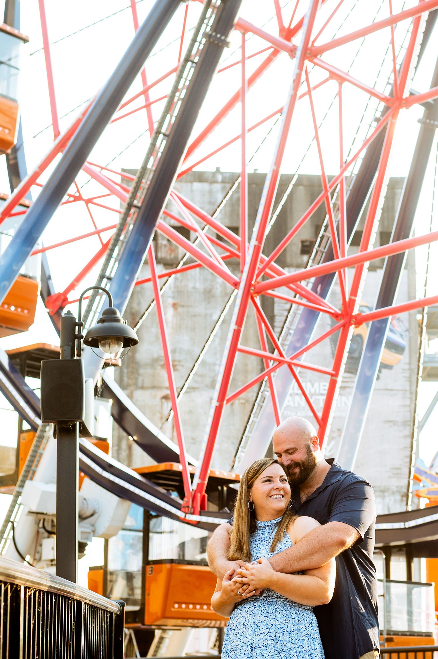 Couple embracing and smiling in front of a colorful Ferris wheel at an amusement park.