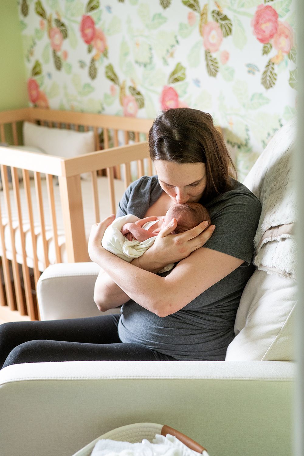 A parent cuddles their newborn in a cozy nursery chair with floral wallpaper behind them.
