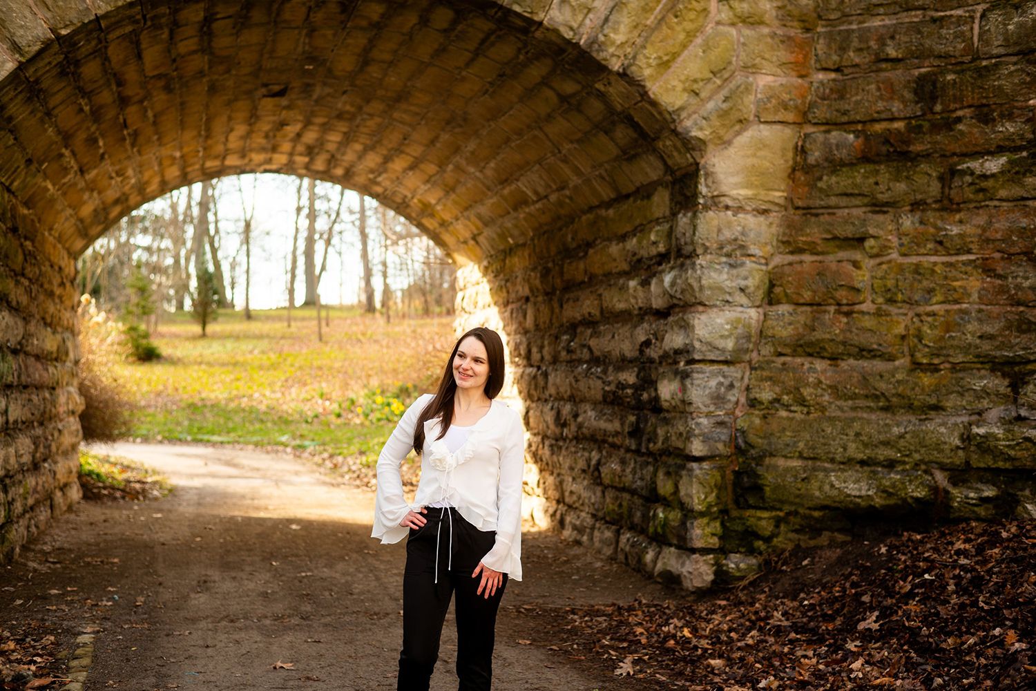 A person in a white top and dark pants poses under a stone arch bridge on an autumn path in a park.