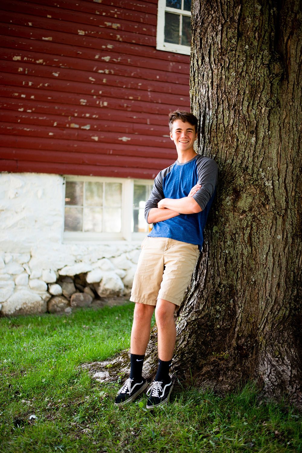 A person in a blue t-shirt and khaki shorts leans against a large tree trunk by a red barn wall.