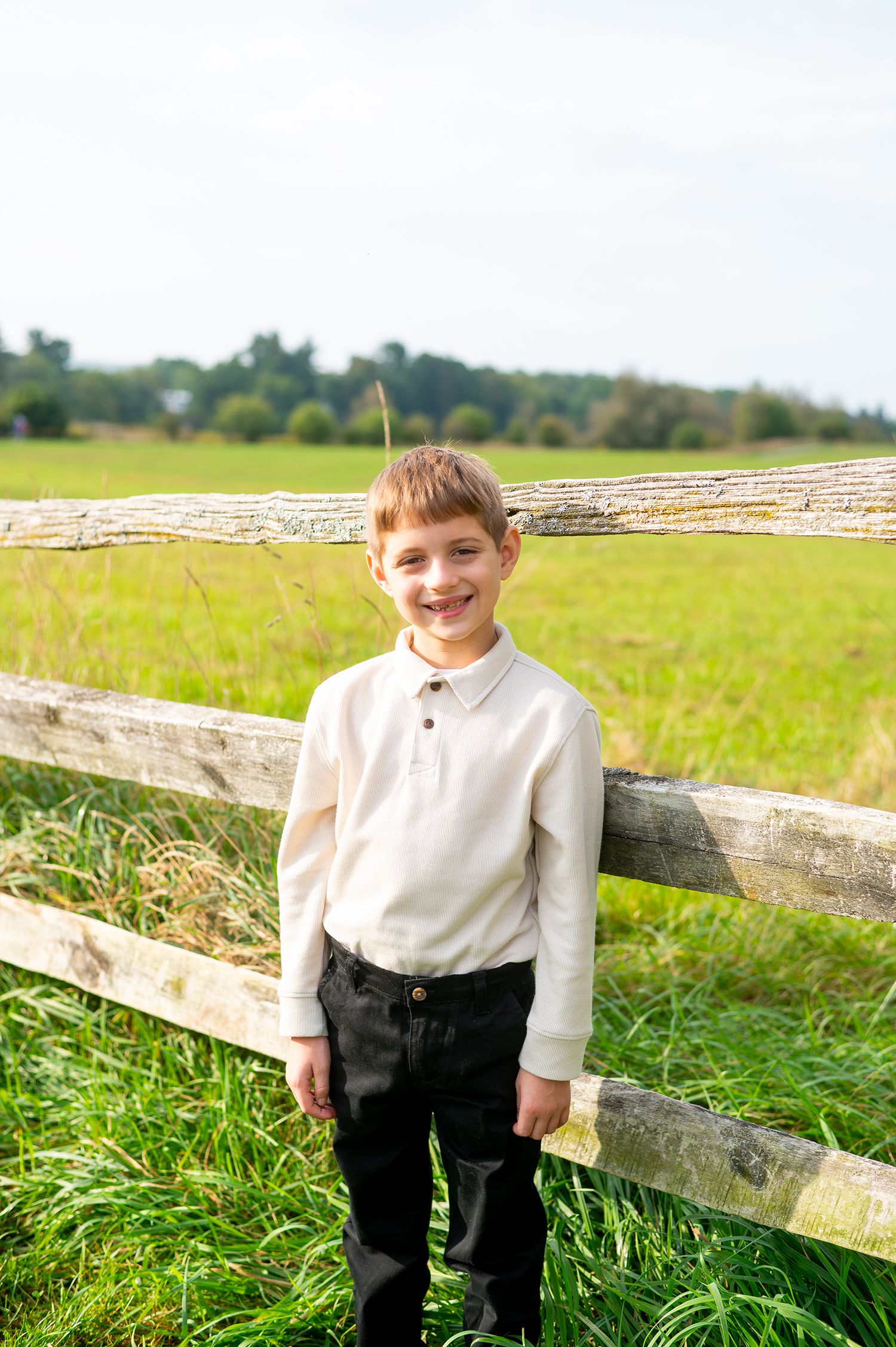 Smiling boy in white shirt and black pants standing by a wooden fence in a green field.