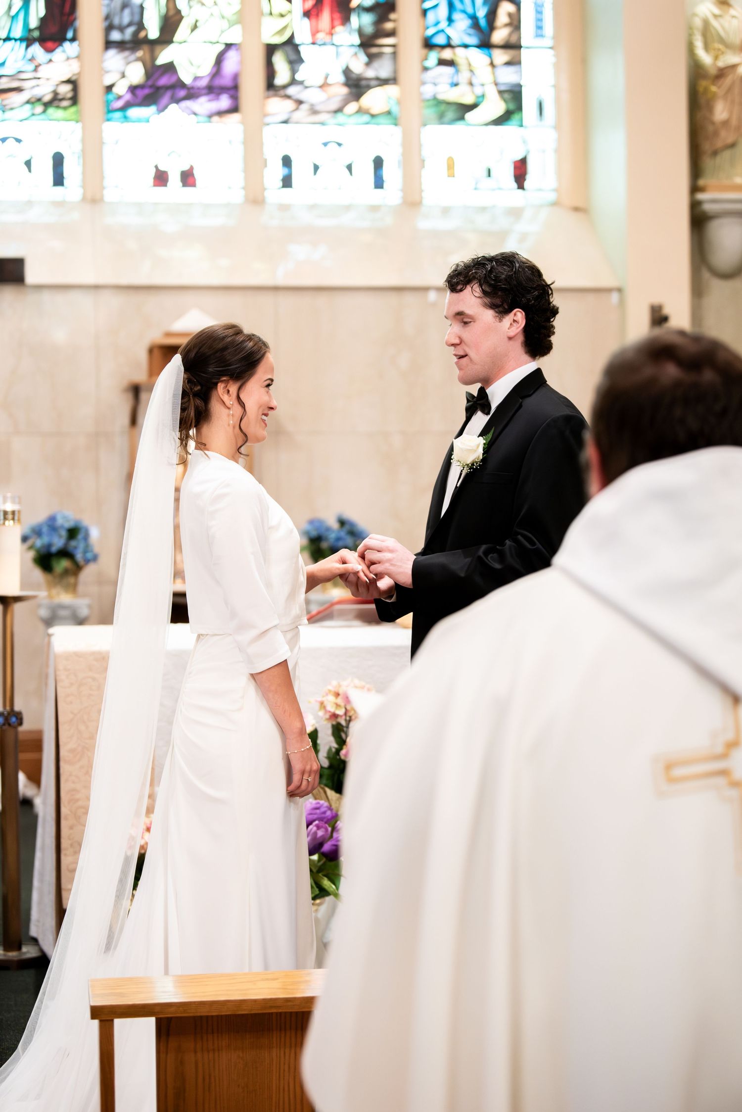A bride and groom exchange vows during their wedding ceremony inside a church with stained glass windows in the background.