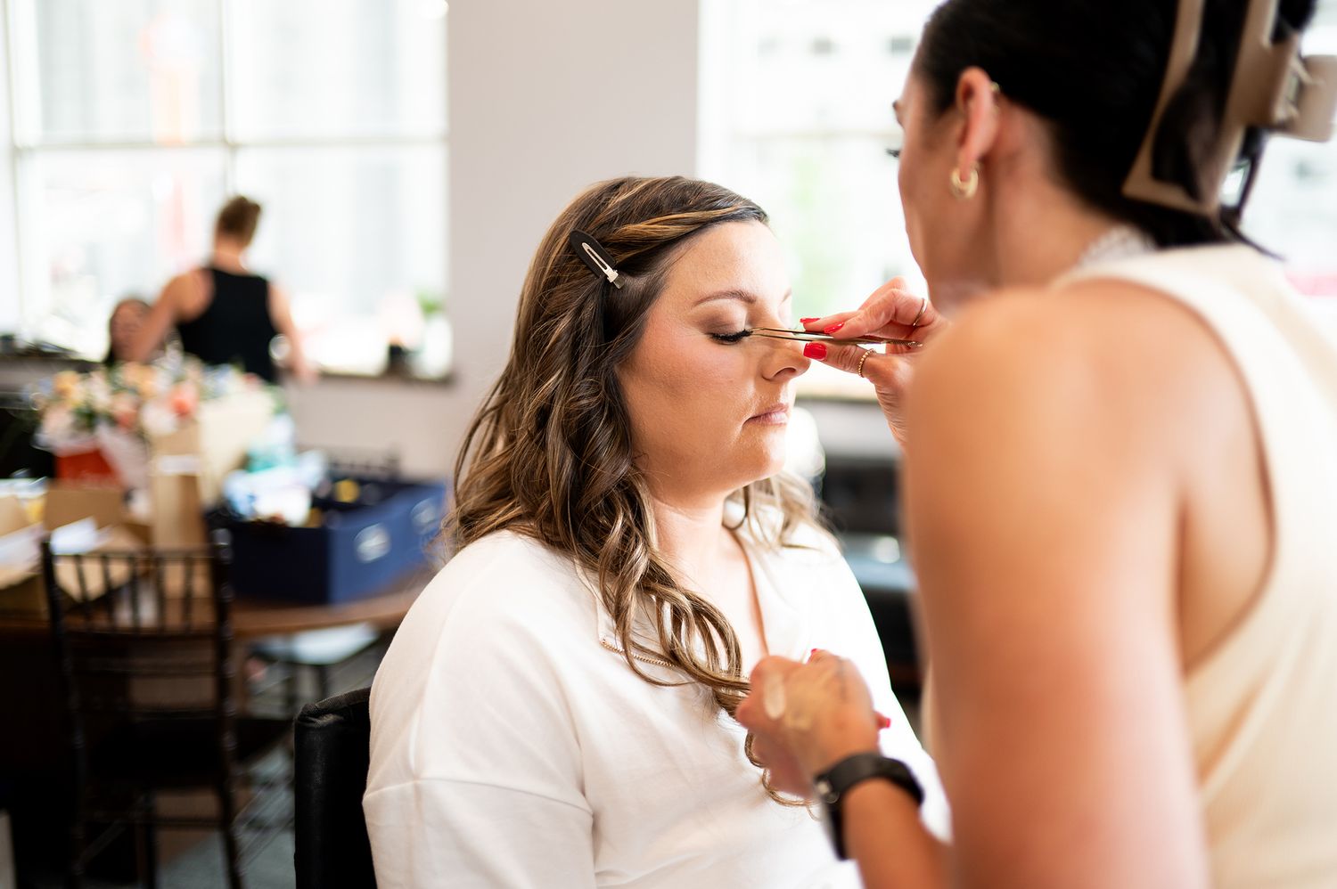 Makeup artist applies cosmetics to a bride's face during wedding preparation while others get ready in the background.