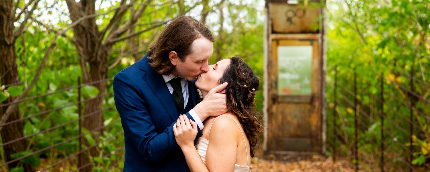 A romantic couple shares an intimate moment outdoors in a lush garden setting with a rustic wooden door in the background.