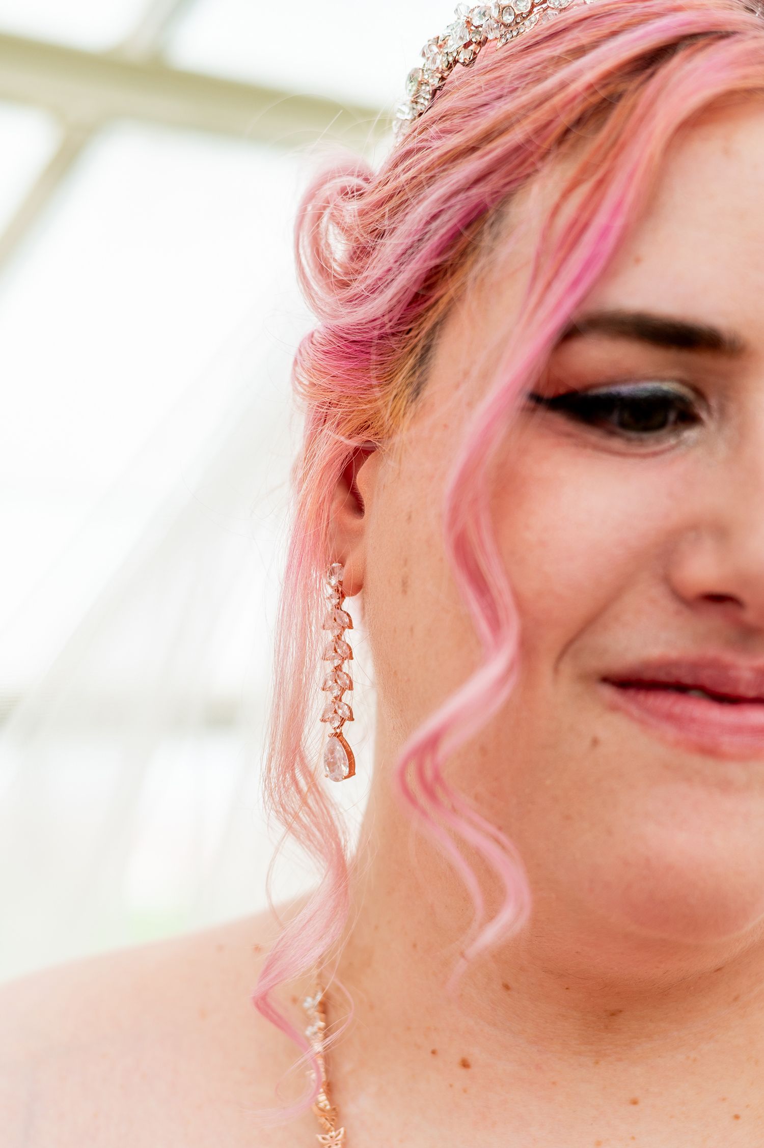 Close-up of a bride with pink curly hair, crystal tiara, and elegant drop earrings, smiling softly.