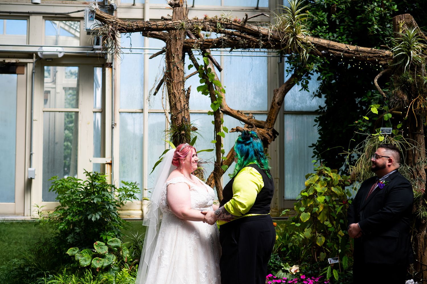 Couple exchanges vows under rustic branch arch in greenhouse wedding ceremony with colorful hair and formal attire.
