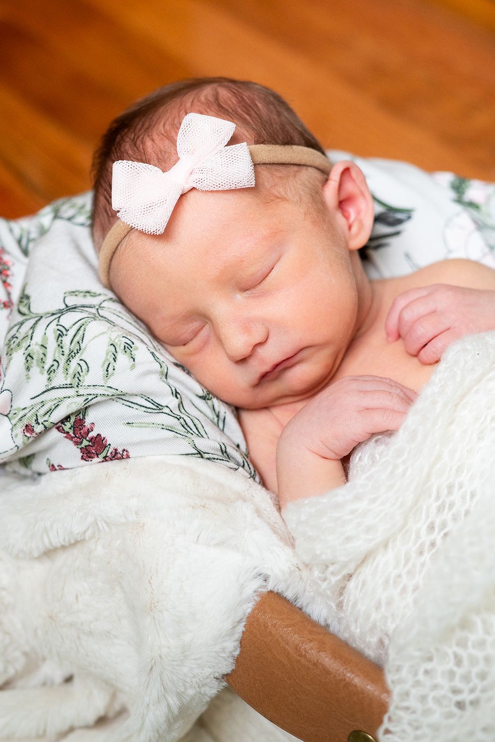 Sleeping newborn baby wearing white bow on head rests peacefully on white lace blanket against floral bedding.