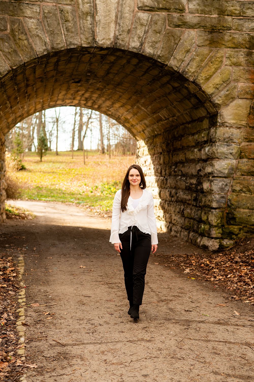 A stone tunnel arch frames a path through autumn trees as someone in a white blouse and dark pants walks through.