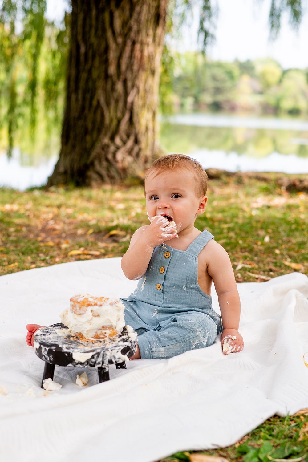 Young child in blue overalls eats cake while sitting on white blanket outdoors near a willow tree.
