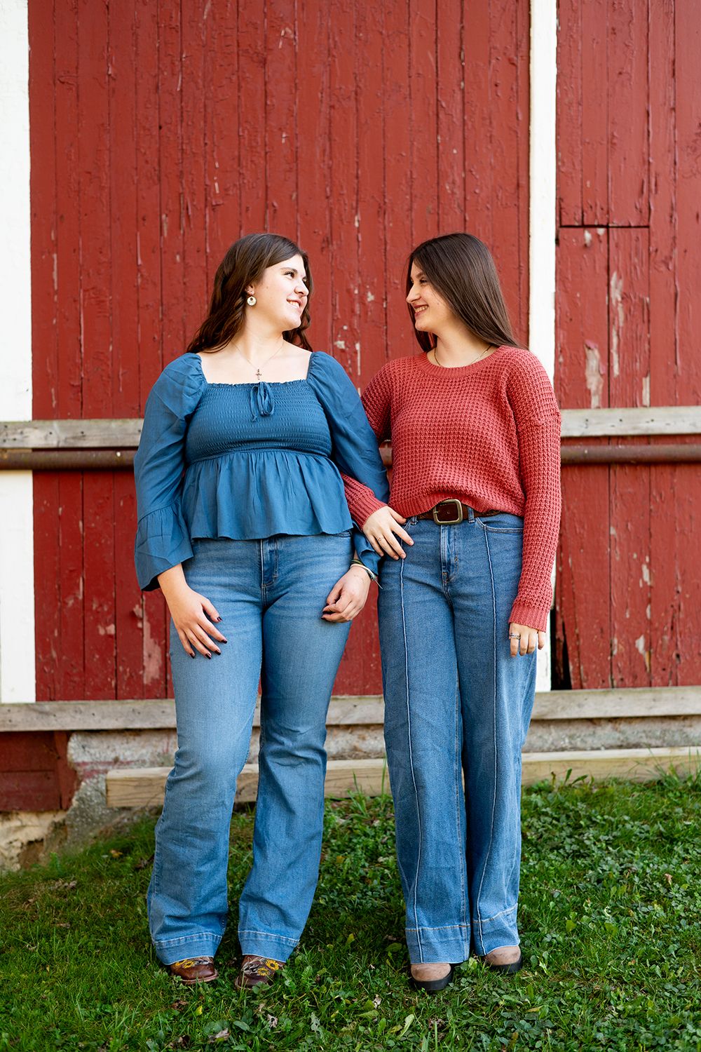 Two women in denim and sweaters stand together in front of a rustic red barn wall on grass.
