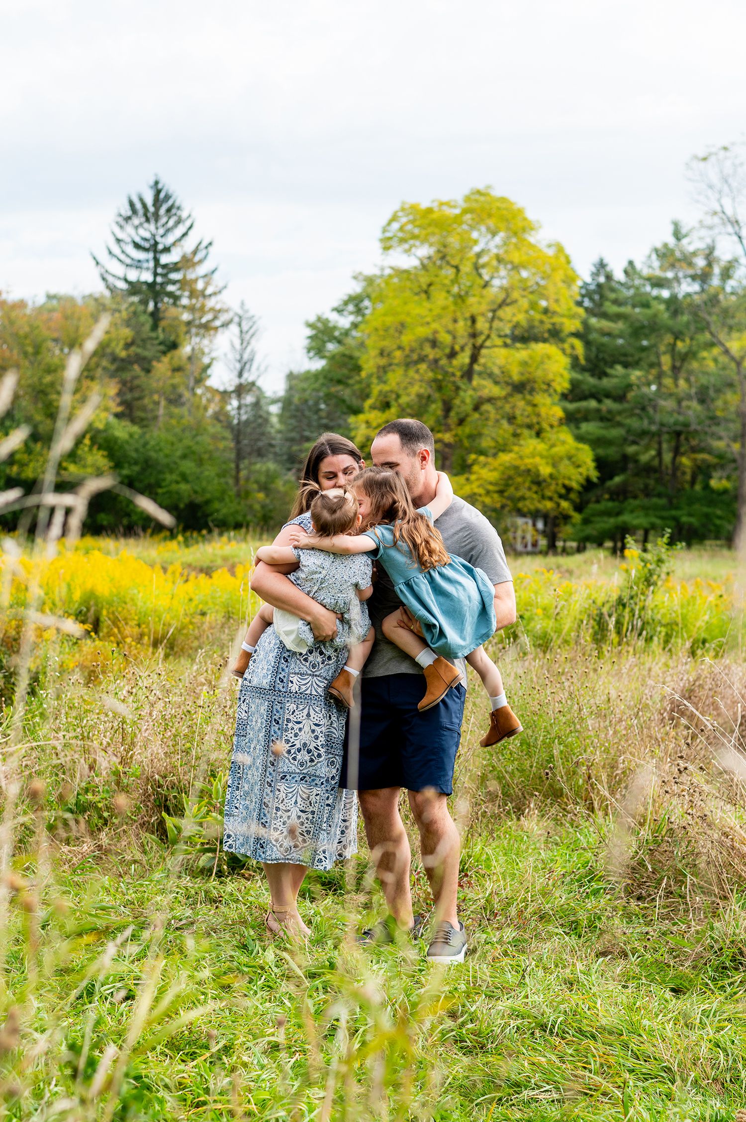Family of four enjoying a fall outdoor photo session in a lush green meadow with colorful autumn trees.