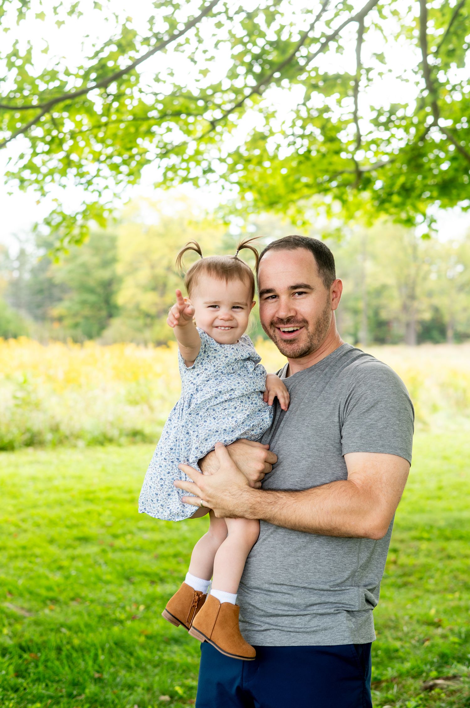 A smiling father holds his cheerful toddler daughter outdoors in a lush green park setting.