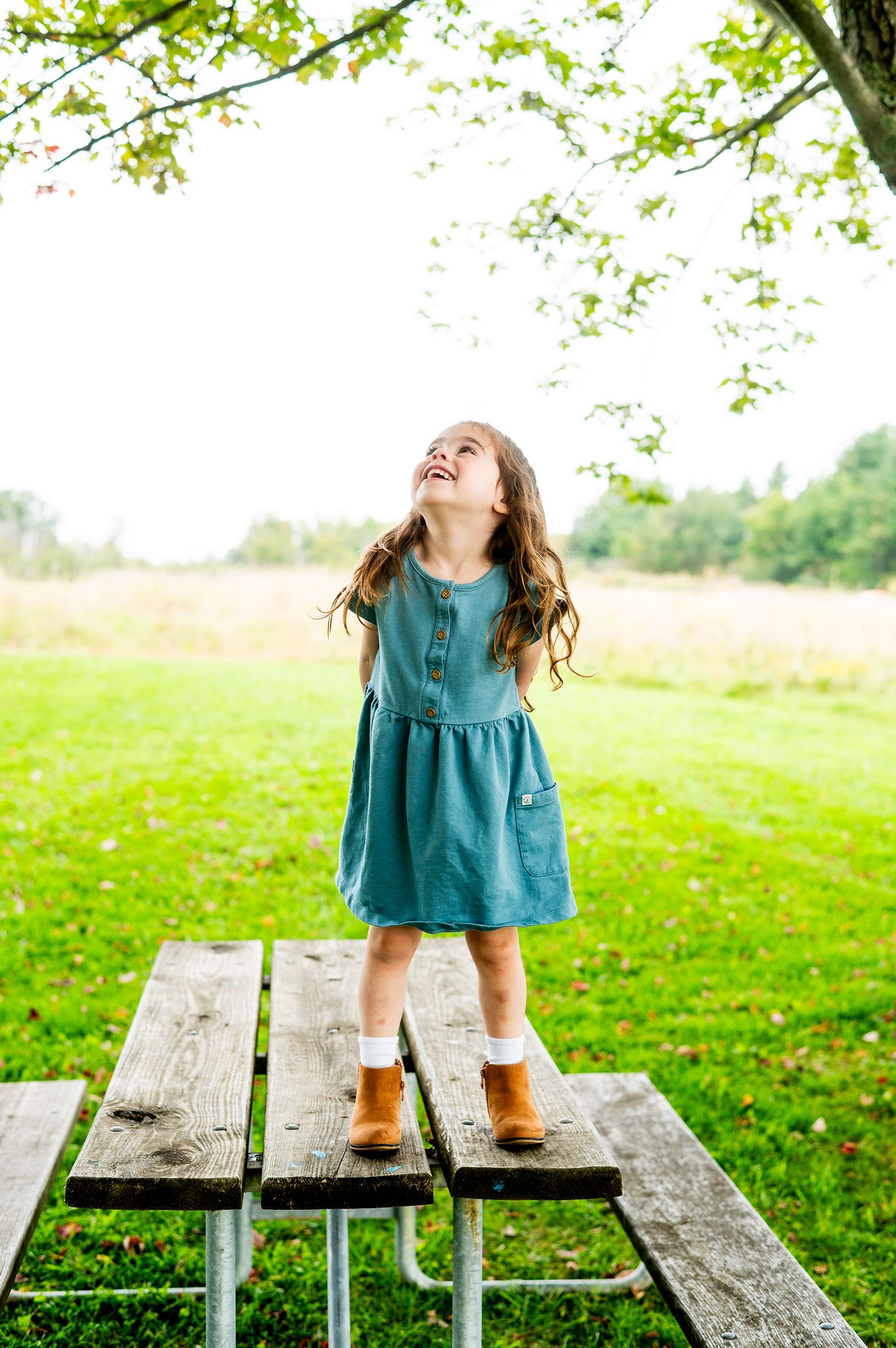 Young girl in blue dress and boots standing on picnic table, laughing and looking up at tree branches.