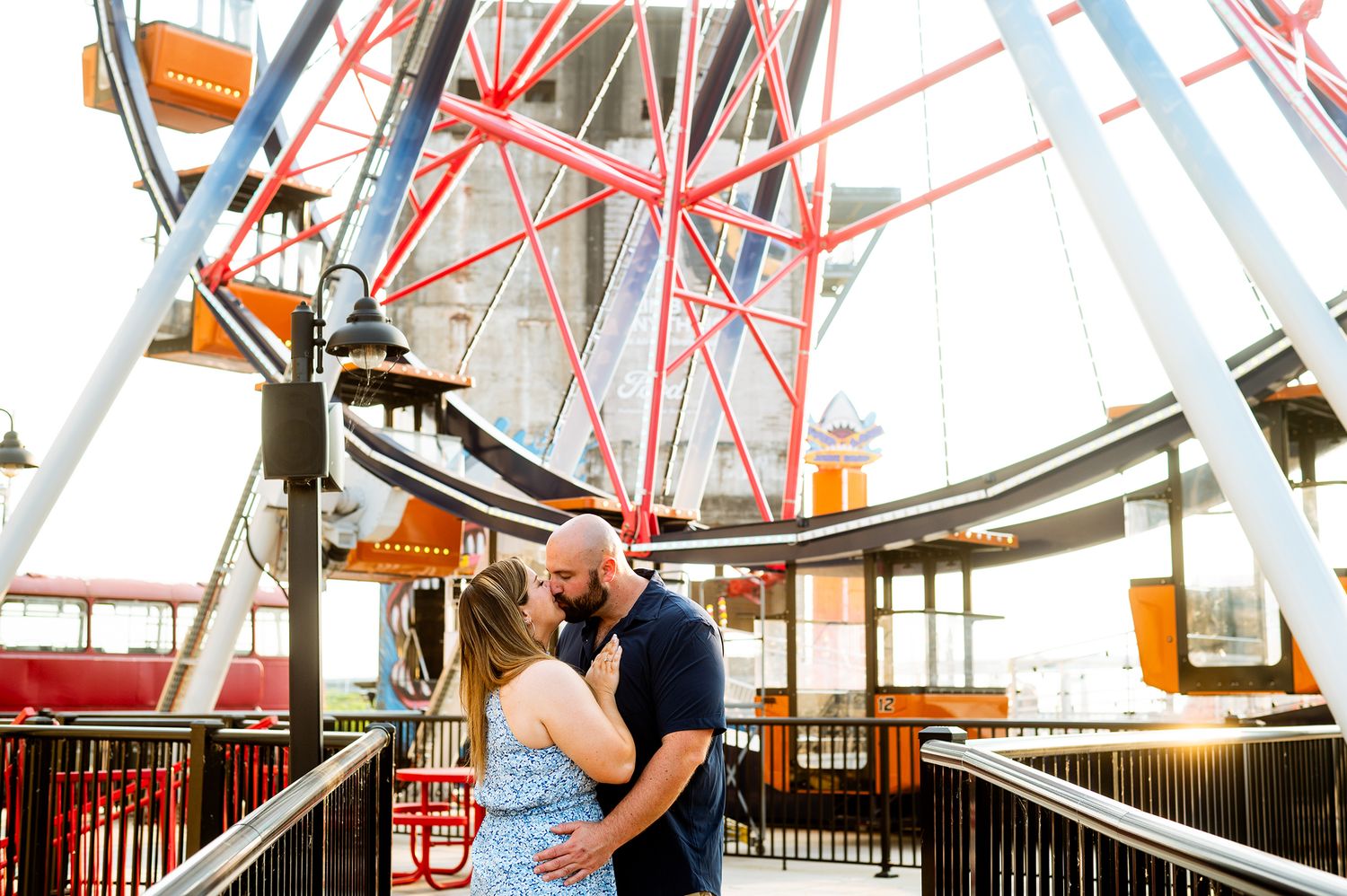 Couple sharing a romantic kiss in front of a colorful Ferris wheel at an amusement park during golden hour.