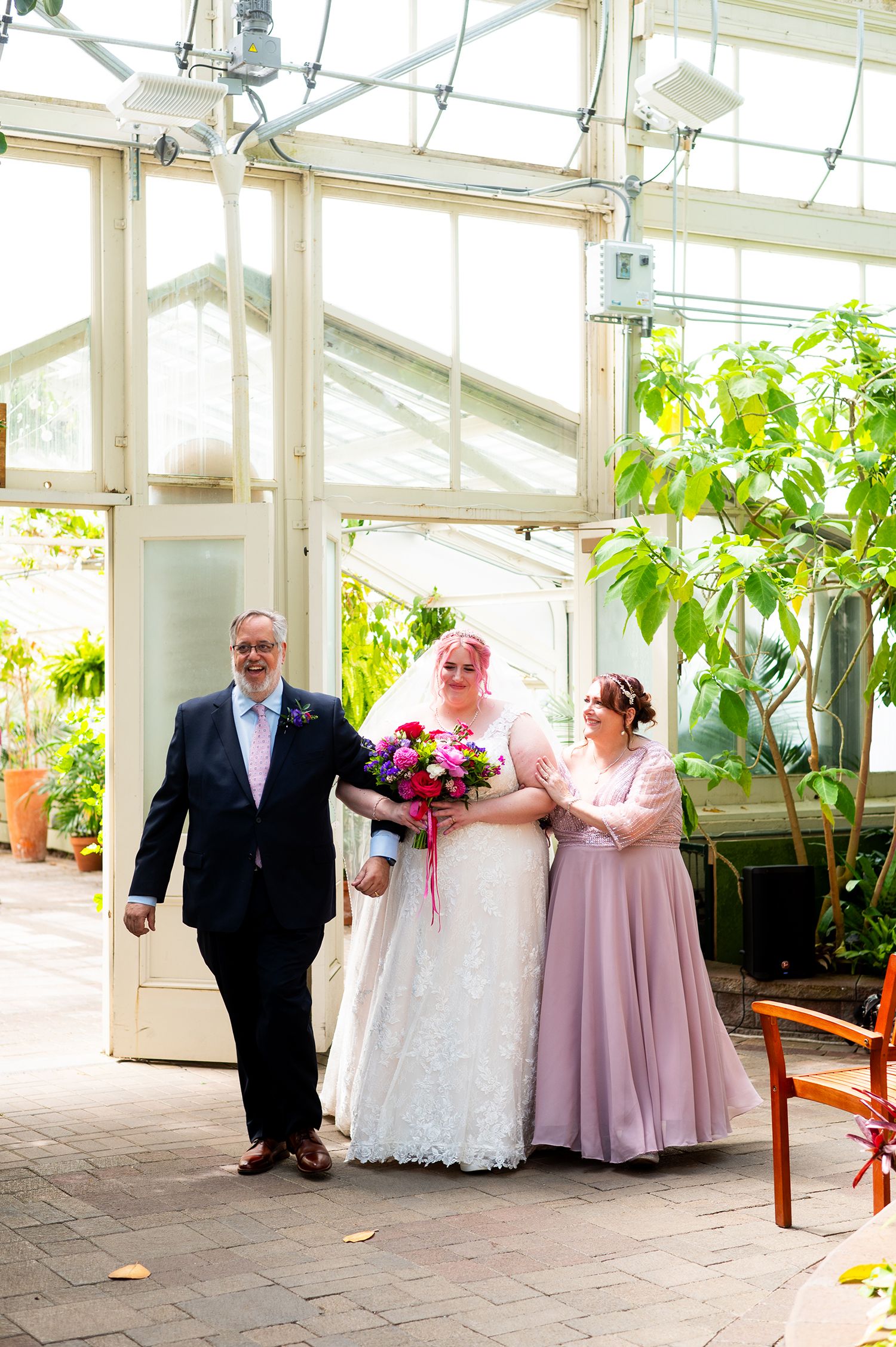 Bride in white lace gown walks with groom in navy suit and bridesmaid in pink dress through a sunlit greenhouse.