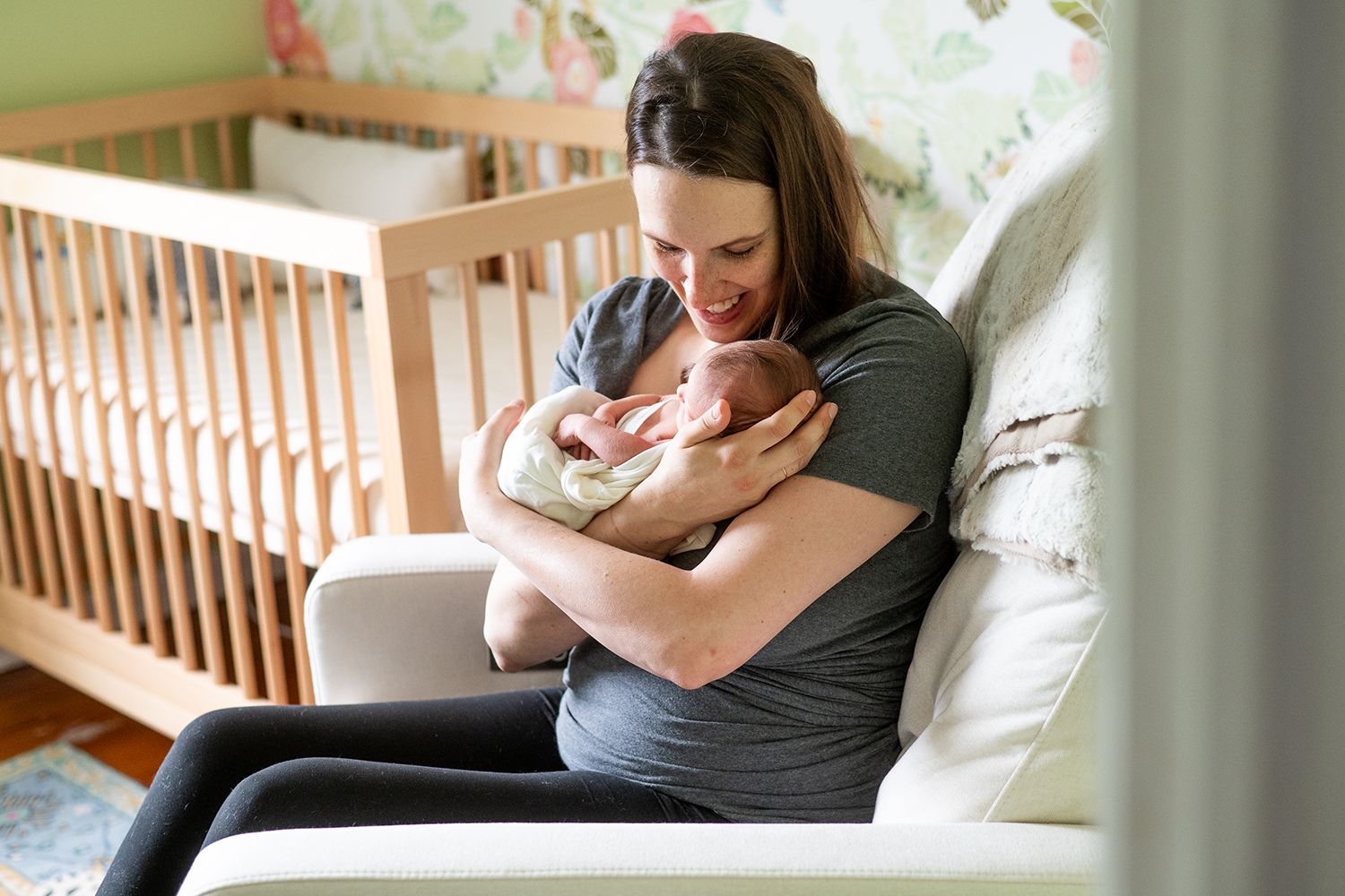 A parent cradles and gazes at their newborn baby while sitting in a rocking chair next to a wooden crib.