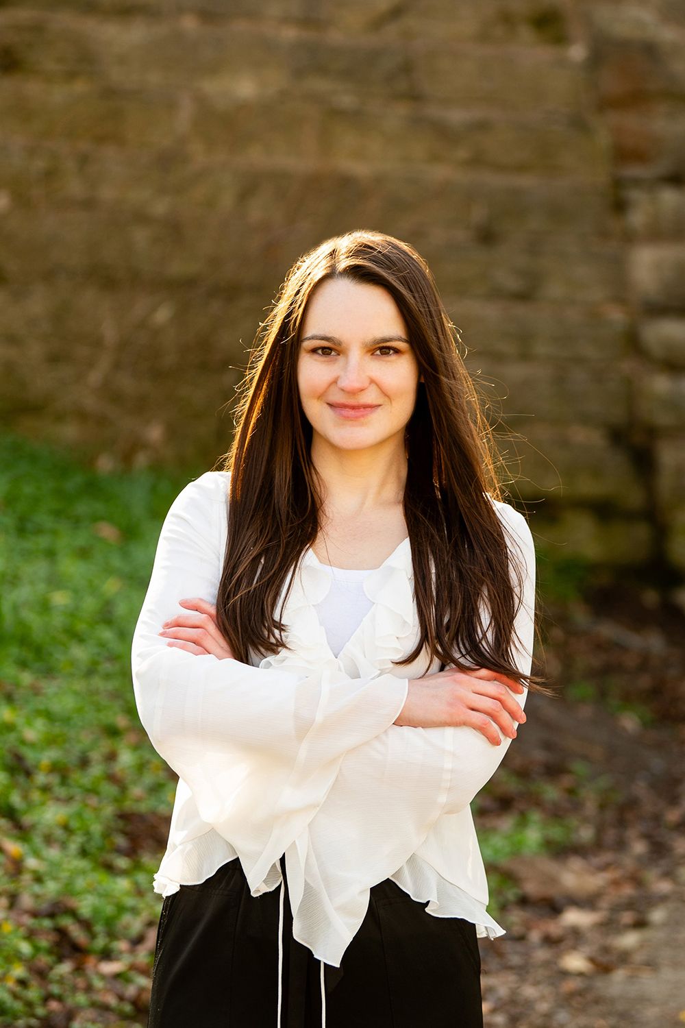 A person in a white flowing top poses with crossed arms against a stone wall background.