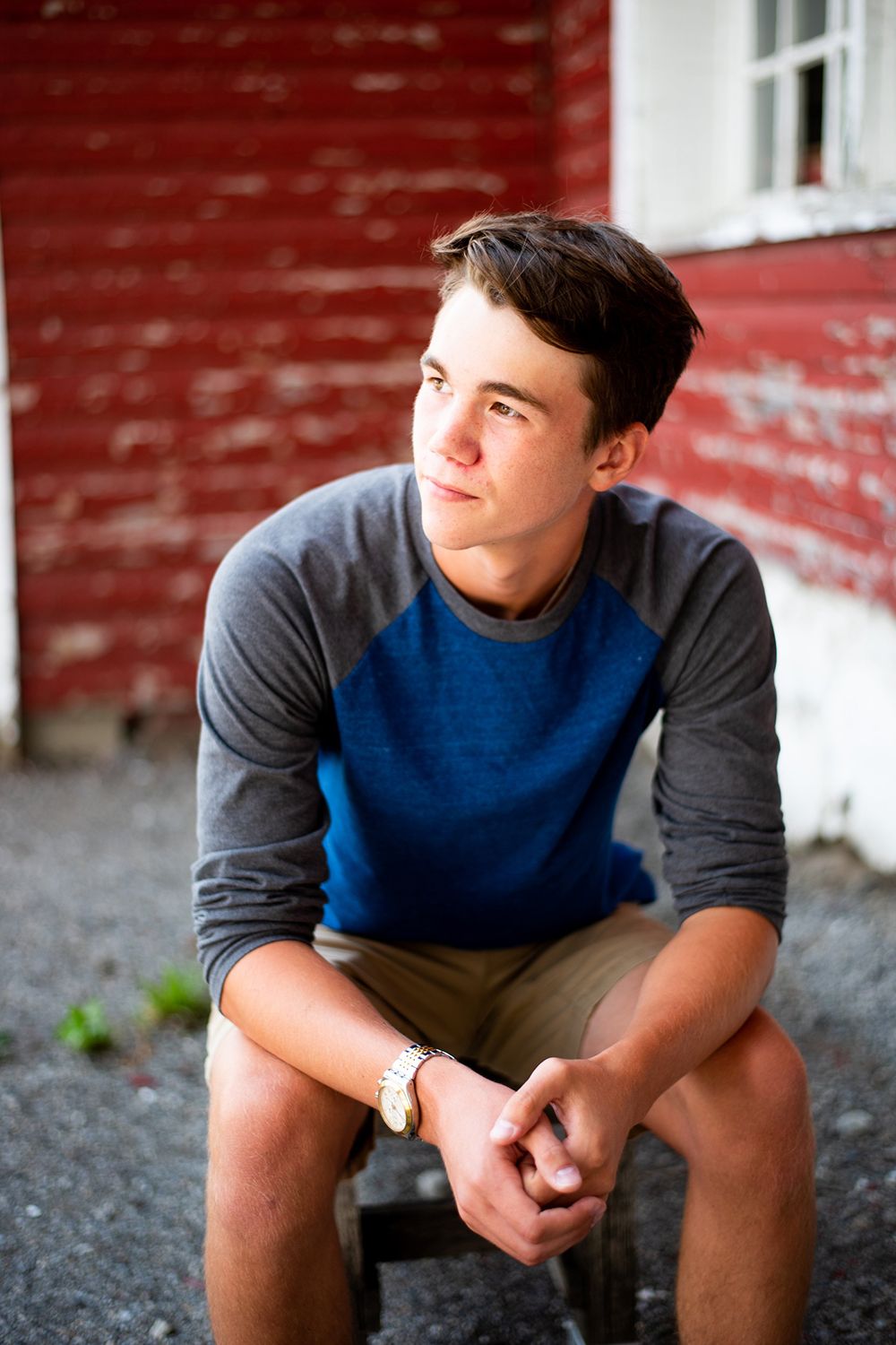 Young person in blue and gray raglan shirt sits thoughtfully on a stool against red barn wall backdrop.