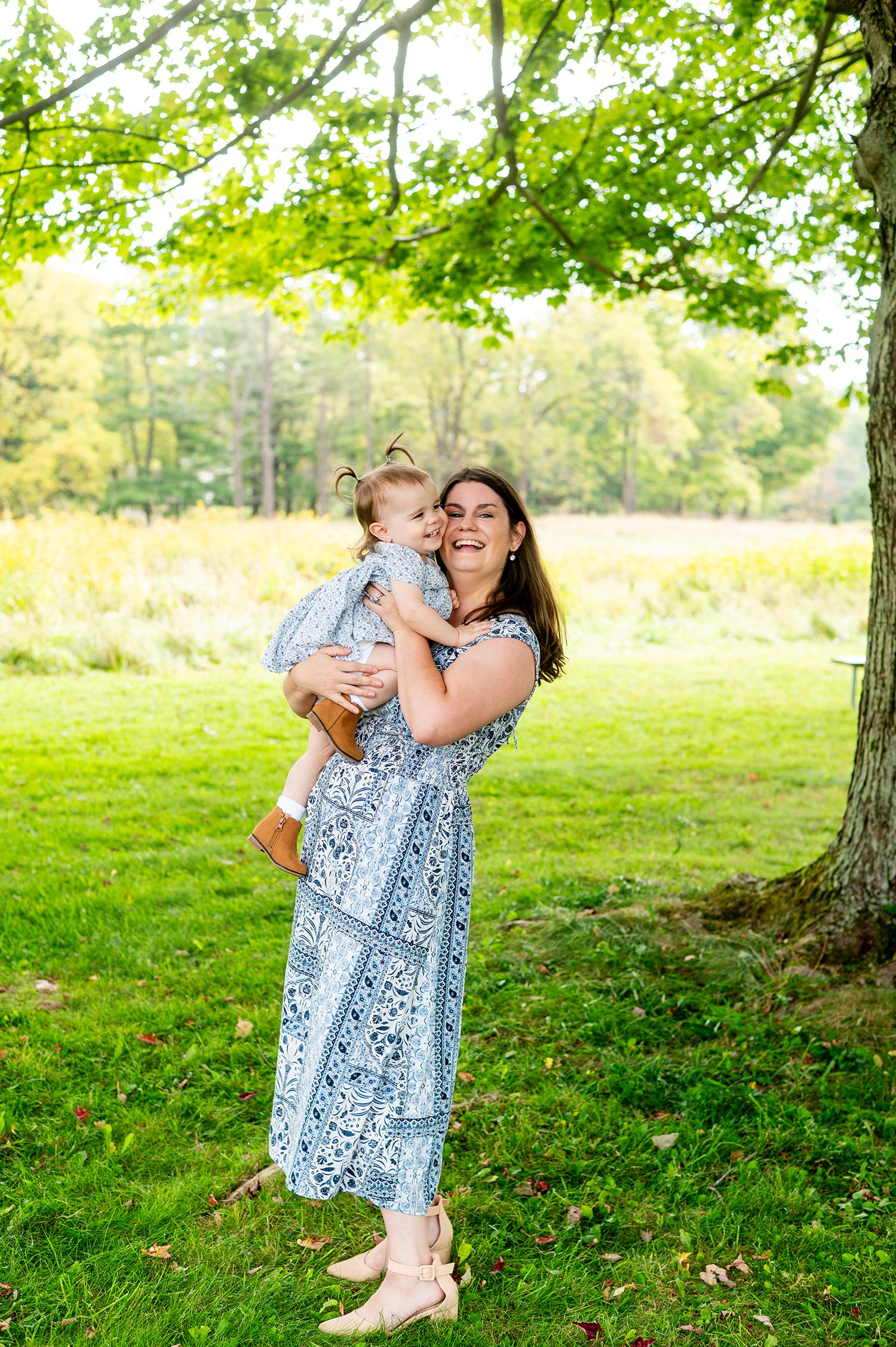 A smiling woman in a blue floral dress holds a baby outdoors in a lush green park setting.