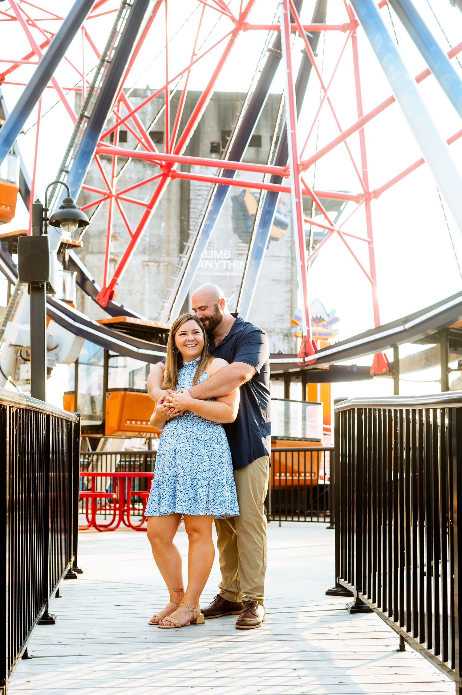 Couple embracing on a boardwalk in front of a colorful Ferris wheel on a sunny day.