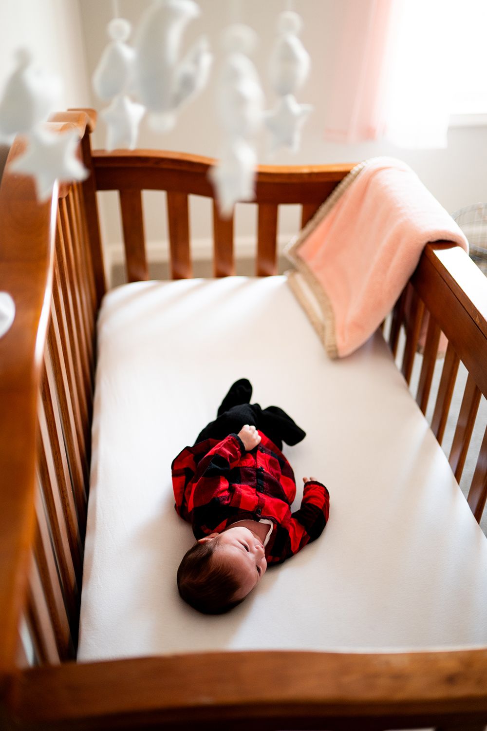 Baby lying in wooden crib wearing red and black plaid outfit with stuffed animal.