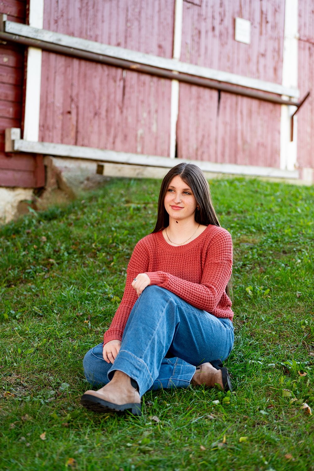 A casual outdoor portrait series of someone in a red sweater and jeans relaxing on green grass near a rustic barn wall.