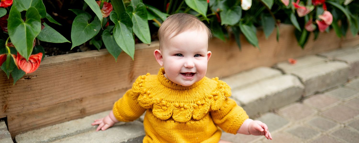 A happy little one in a golden yellow ruffled sweater sits near a planter with red flowers and green foliage.