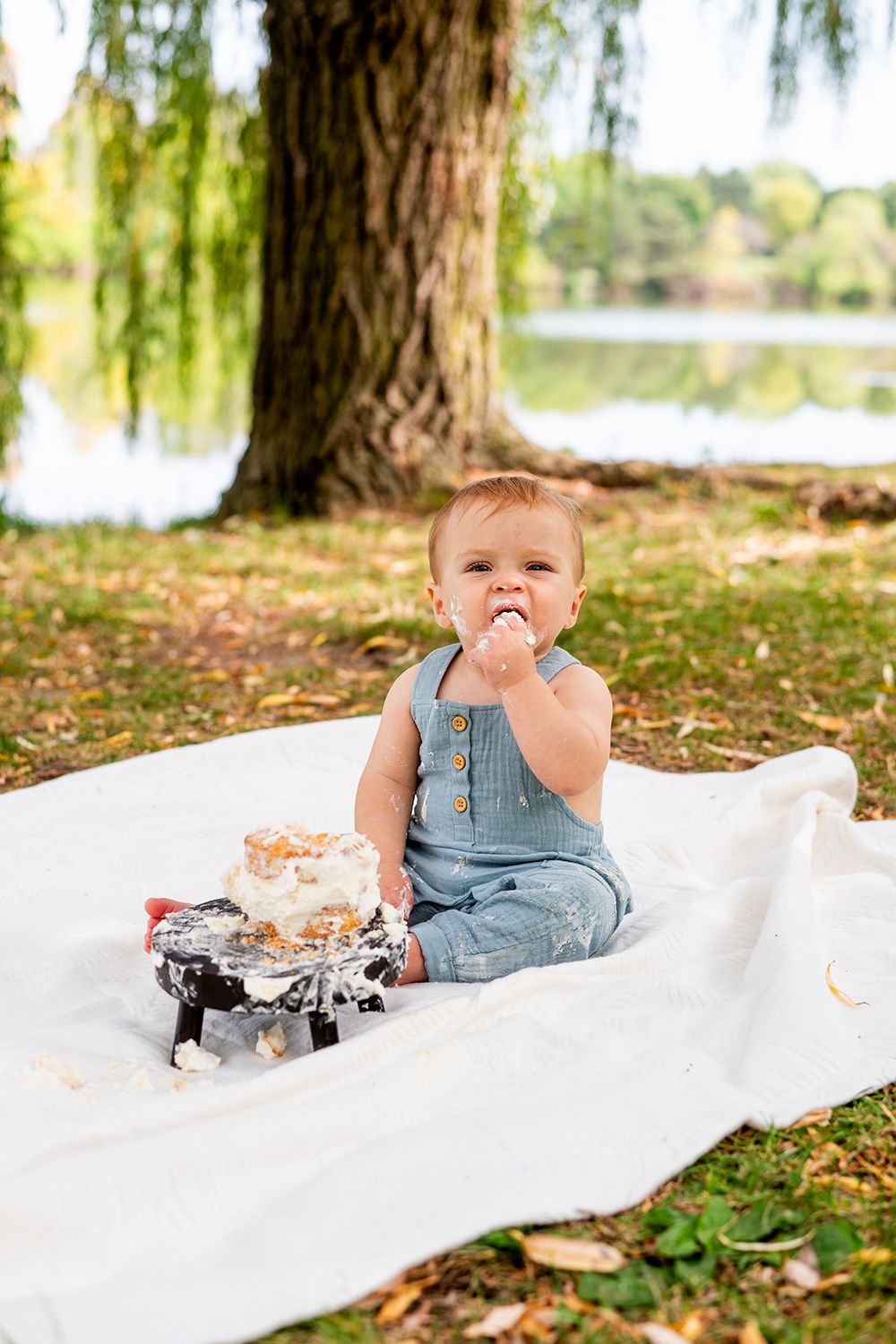 A happy toddler in blue overalls sits on a white blanket outdoors eating cake near a lake.